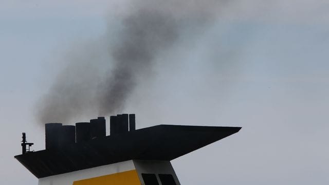 Eine dunkle Abgaswolke steigt aus dem Schornstein eines Fährschiffs bei der Ausfahrt aus dem Seehafen.