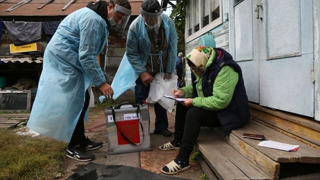 Zwei Wahlhelfer stehen neben einer Frau, die auf den Stufen vor einem Haus sitzt und Dokumente ausfüllt.