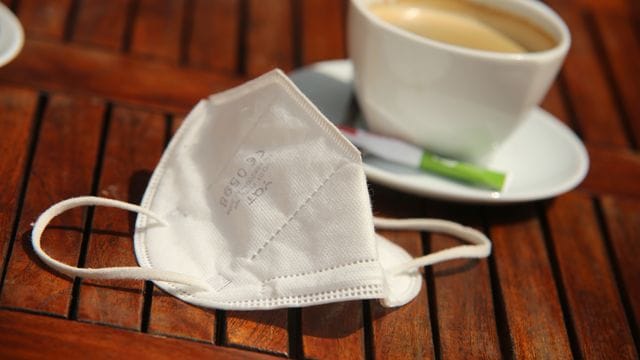 Eine FFP2-Maske liegt neben einer Tasse Kaffee auf einem Tisch.