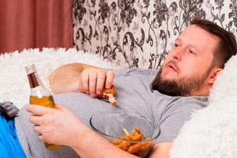 Ein Mann liegt im Bett, trinkt ein Bier und isst: Zu viel Bequemlichkeit ist ungesund und begünstigt zahlreiche Krankheiten.