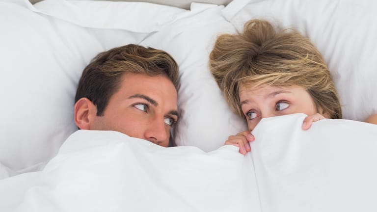 Ein Paar liegt im Bett und schaut sich besorgt an. Bakterielle Infektionen im Intimbereich können durch Geschlechtsverkehr an den Partner weitergegeben werden.