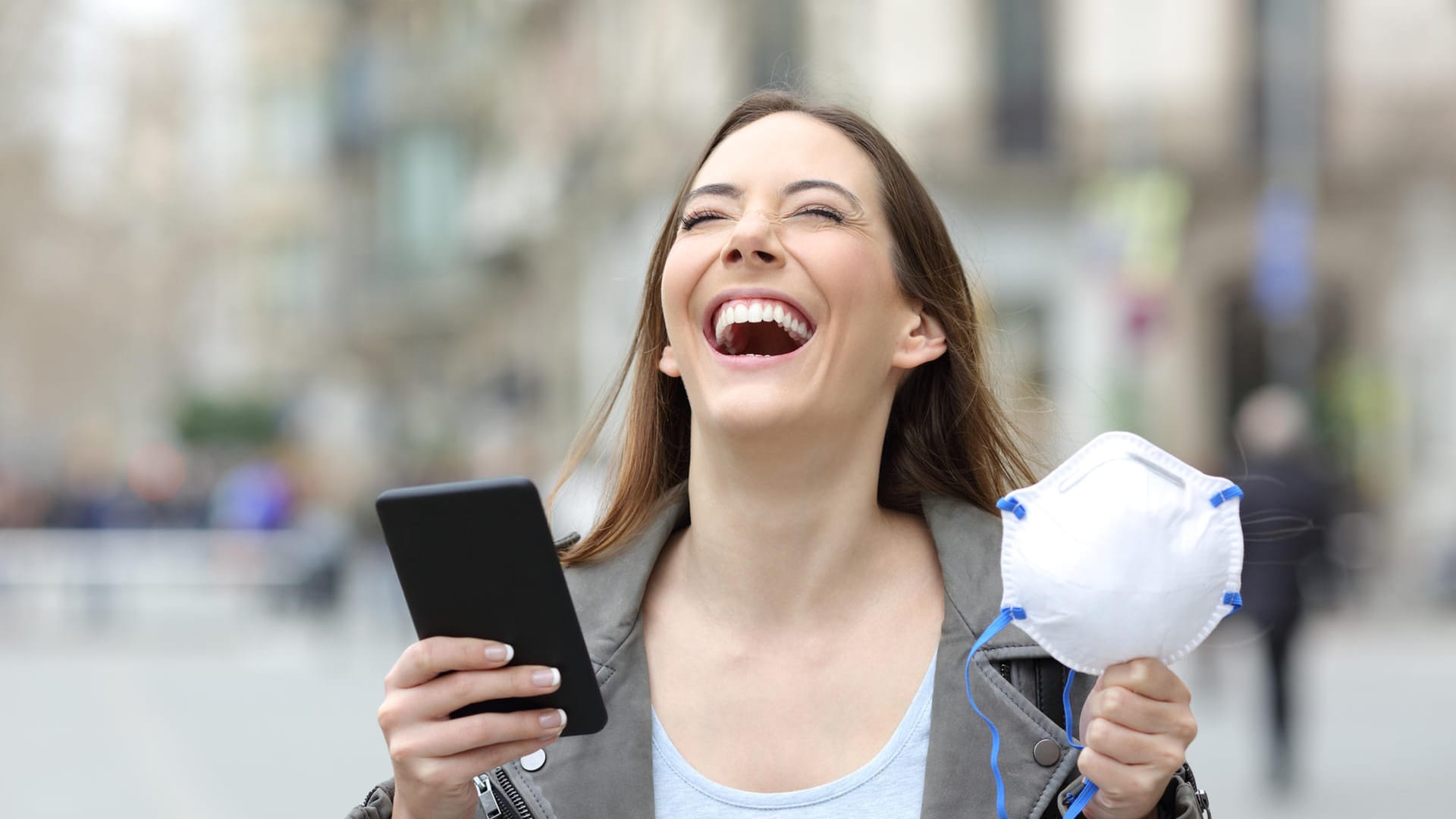 Eine lachende Frau mit Handy, die ihre Maske ausgezogen hat. In stressigen Phasen wie der Corona-Pandemie ist Humor Gold wert.