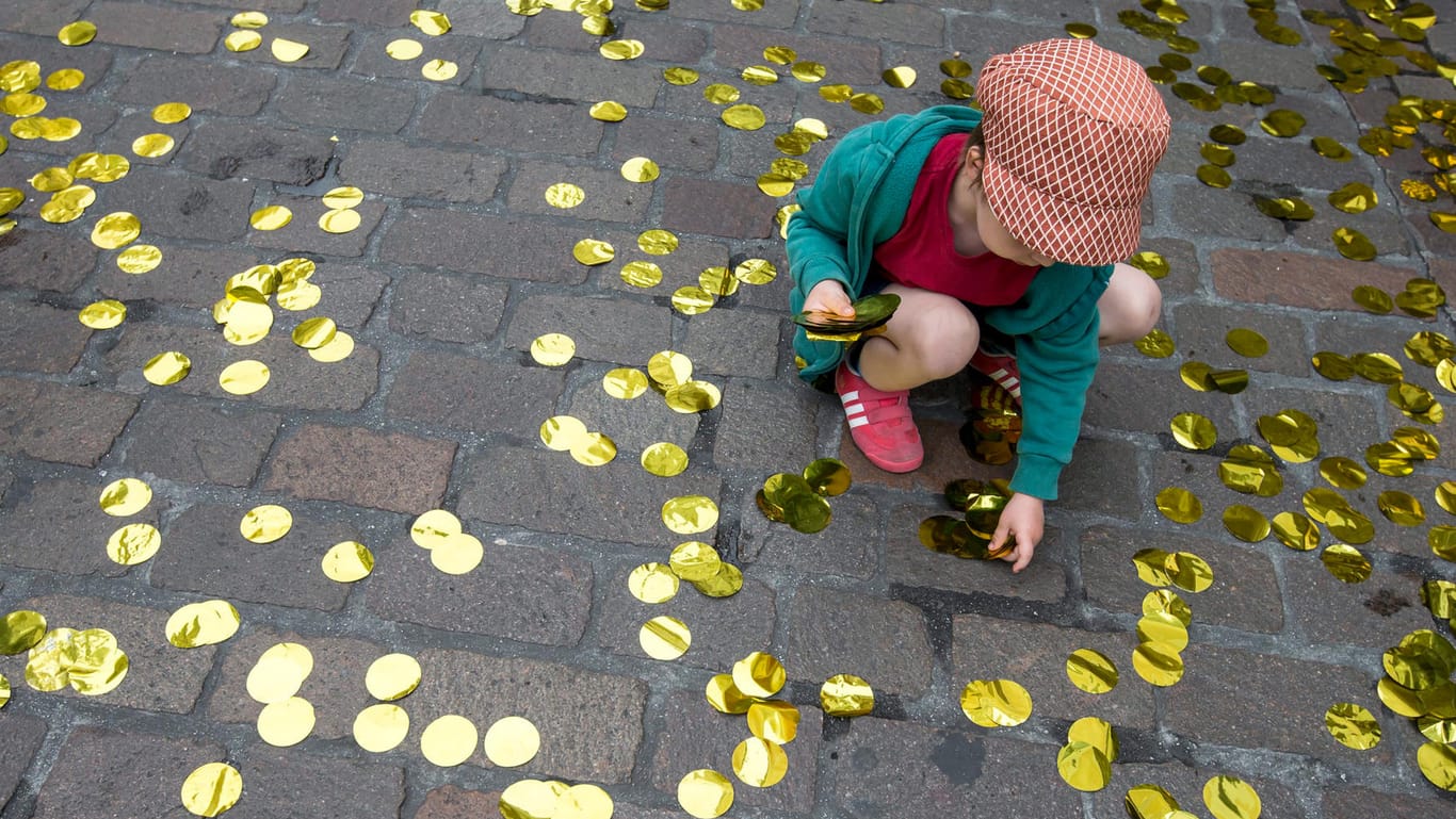 Ein kleines Mädchen sammelt bei einer Veranstaltung zum bedingungslosen Grundeinkommen goldene Konfetti-Taler von der Straße auf.