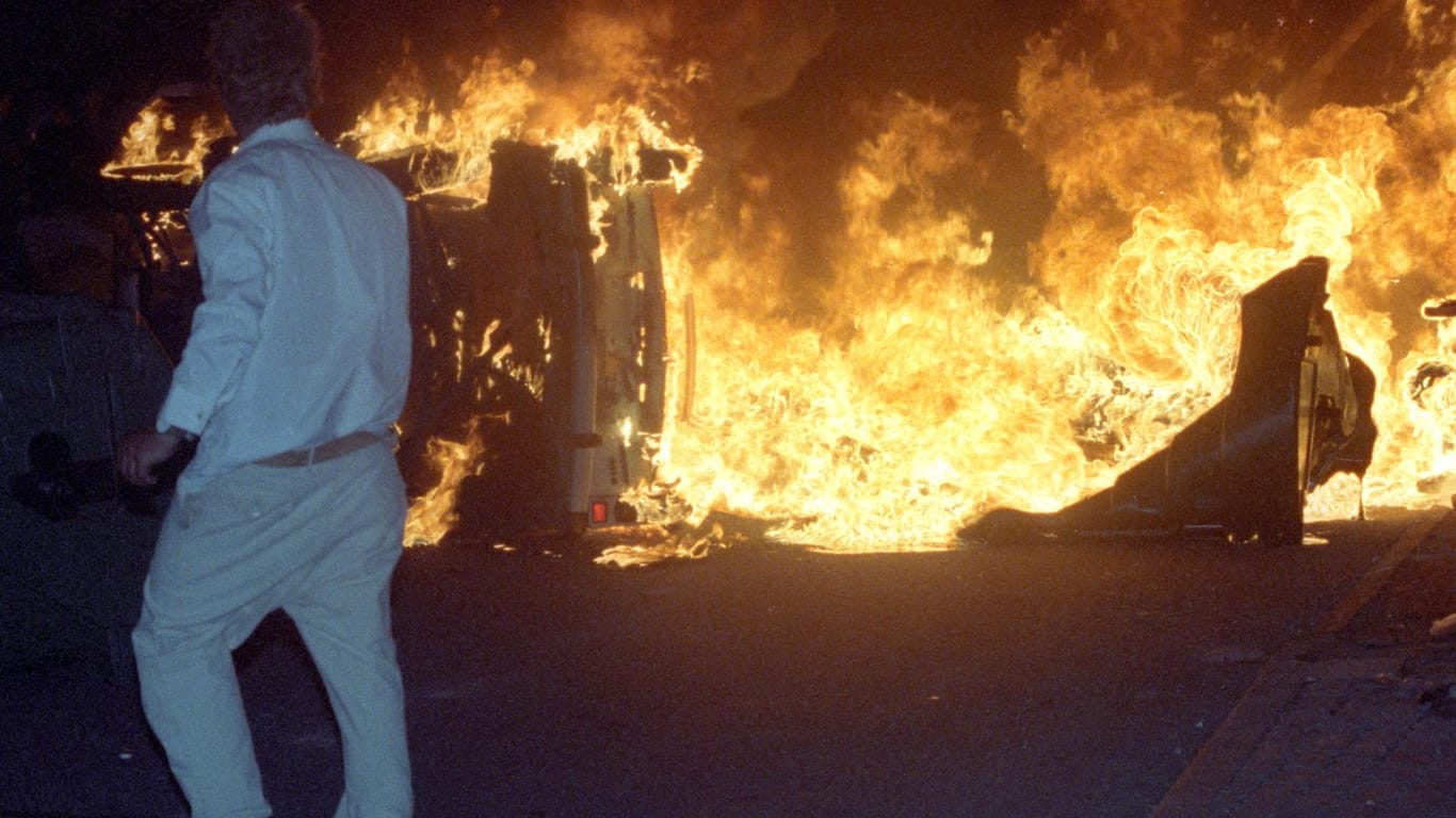 Ein Mann steht 1992 vor einem brennenden Pkw auf einer Straße am zentralen Asylbewerberheim von Mecklenburg-Vorpommern in Rostock-Lichtenhagen.
