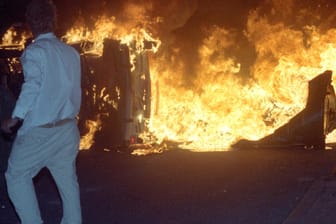 Ein Mann steht 1992 vor einem brennenden Pkw auf einer Straße am zentralen Asylbewerberheim von Mecklenburg-Vorpommern in Rostock-Lichtenhagen.