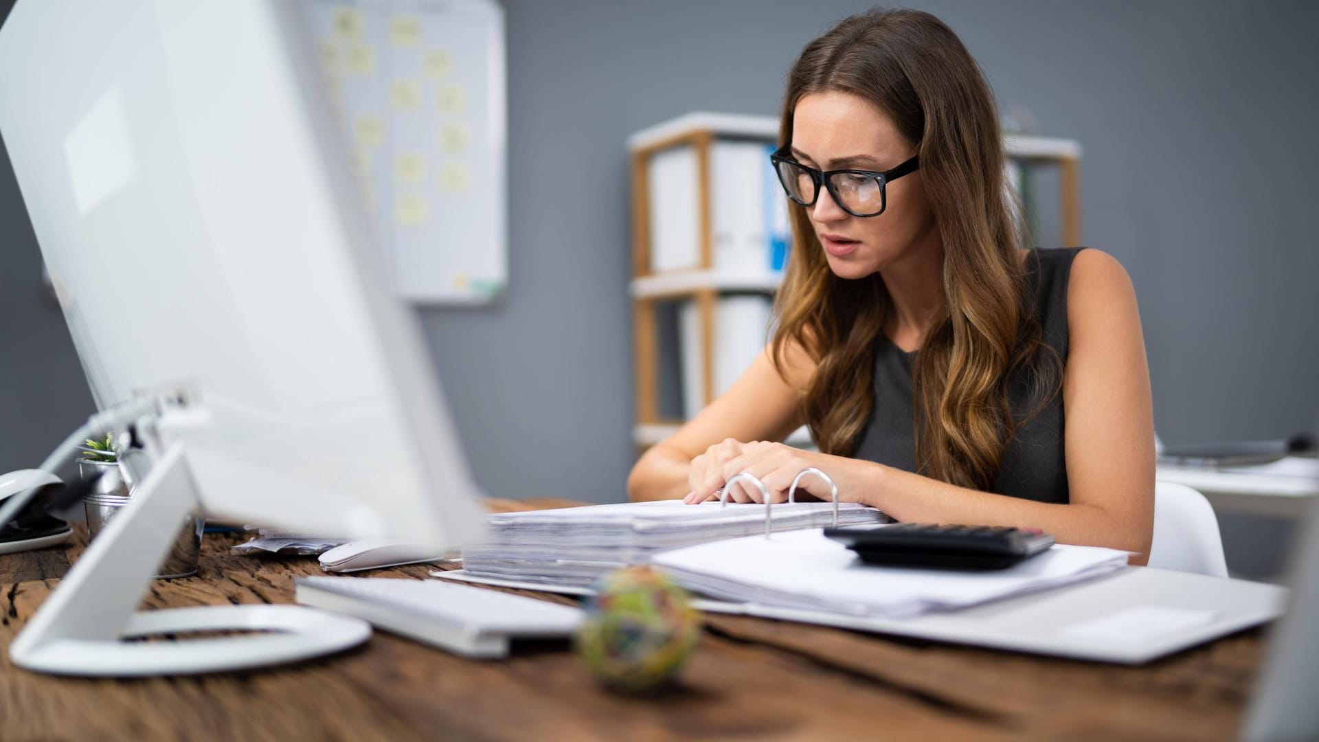 Brünette Frau mit Brille erledigt bürokratische Aufgaben an einem Computer mit einem offenen Ordner.