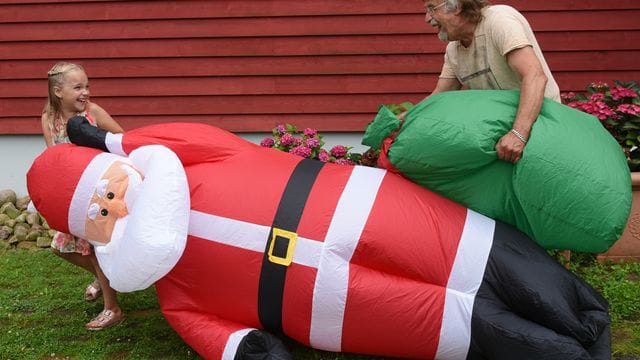 Lachend tragen Alicia und ihr Opa Dietmar Sawatzke den drei Meter großen aufgeblasenen Weihnachtsmann neben die mit Sommerblumen dekorierte Eingangstür.