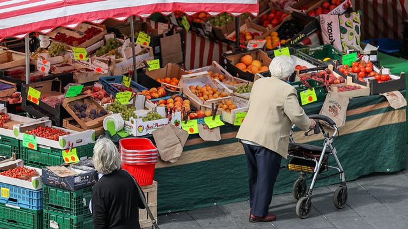 Eine Seniorin steht, gestützt auf ihren Rollator, vor einem Obst- und Gemüsestand auf einem Wochenmarkt.