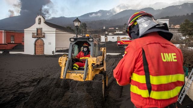 Einsatzkräfte des Militärs entfernen schwarze Asche vom Vulkan, der hinter der kleinen Kirche auf der Kanareninsel La Palma weiter Lava ausstößt. Einsatzkräfte des Militärs entfernen schwarze Asche vom Vulkan, der hinter der kleinen Kirche auf der Kanareninsel La Palma weiter Lava ausstößt.
