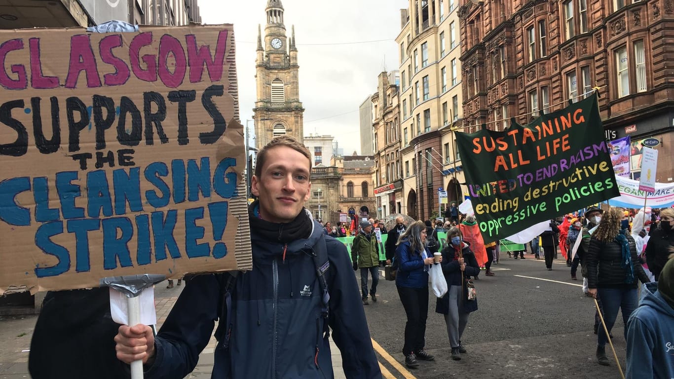 Liams Plakat hat auf jeder Seite eine andere Aufschrift: Die eine fordert stärkeren Klimaschutz, auf der anderen solidarisiert er sich mit den Müllwerkern in Glasgow, die bessere Arbeitsbedingungen fordern.