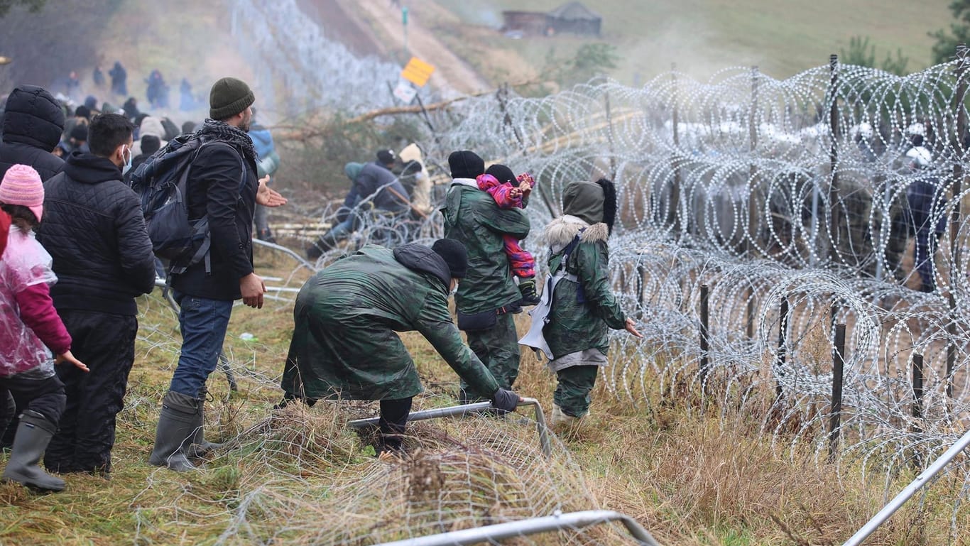 Migranten stoßen an der belarussischen-polnischen Grenze einen Zaun um, während die Polizei hinter einem Stacheldrahtzaun steht.