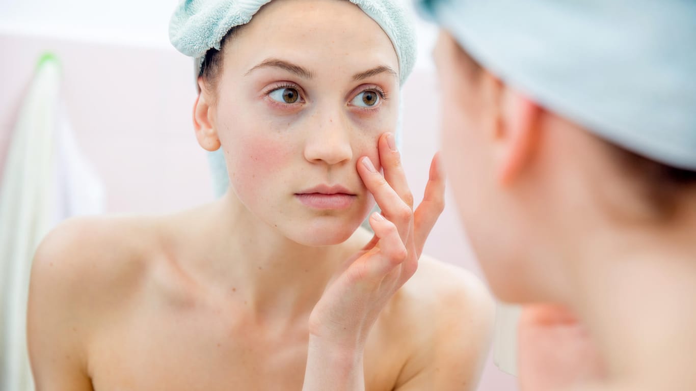 Frau schaut in den Spiegel: Ausgeprägter Eisenmangel kann zu blasser Haut führen – doch nicht alle Symptome sind so offensichtlich.