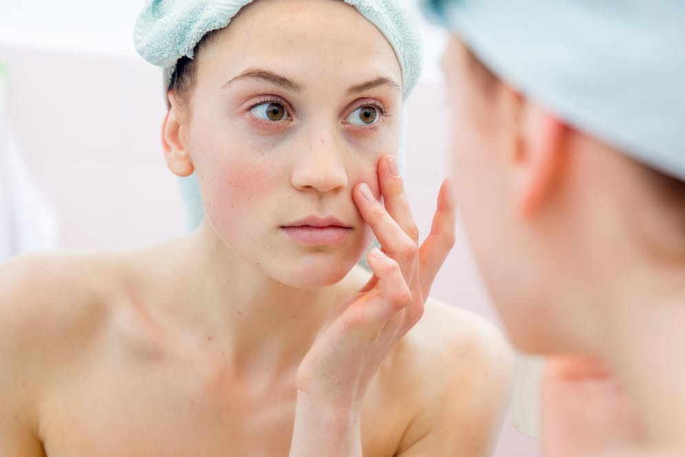 Frau schaut in den Spiegel: Ausgeprägter Eisenmangel kann zu blasser Haut führen – doch nicht alle Symptome sind so offensichtlich.