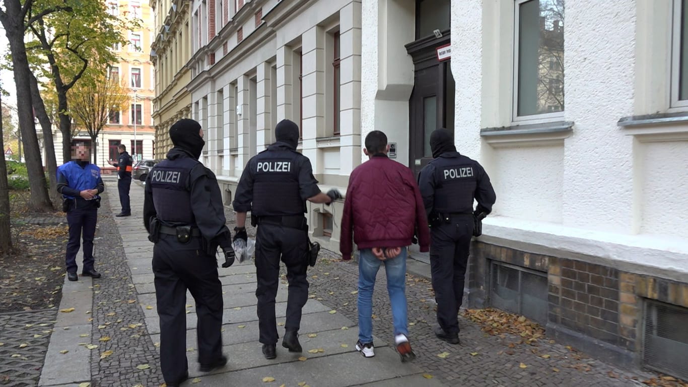 Eine Person in Handschellen läuft neben Polizeibeamten: In Leipzig haben hunderte Beamte Durchsuchungen wegen gefälschter Pässe durchgeführt.