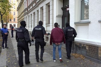 Eine Person in Handschellen läuft neben Polizeibeamten: In Leipzig haben hunderte Beamte Durchsuchungen wegen gefälschter Pässe durchgeführt.