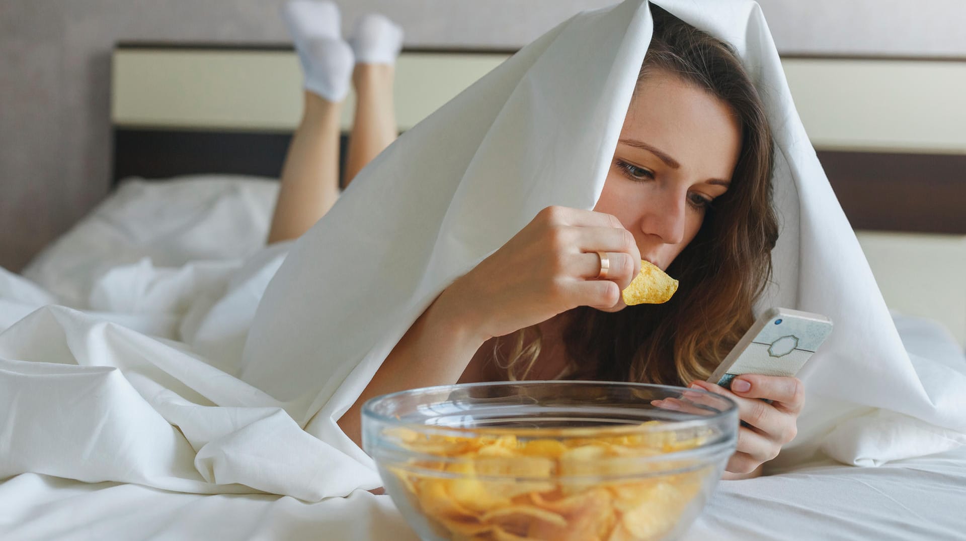 Eine junge Frau isst Chips im Bett und schaut dabei auf ihr Handy. Falsche Ernährungsgewohnheiten können dazu führen, dass man schlechter schläft.