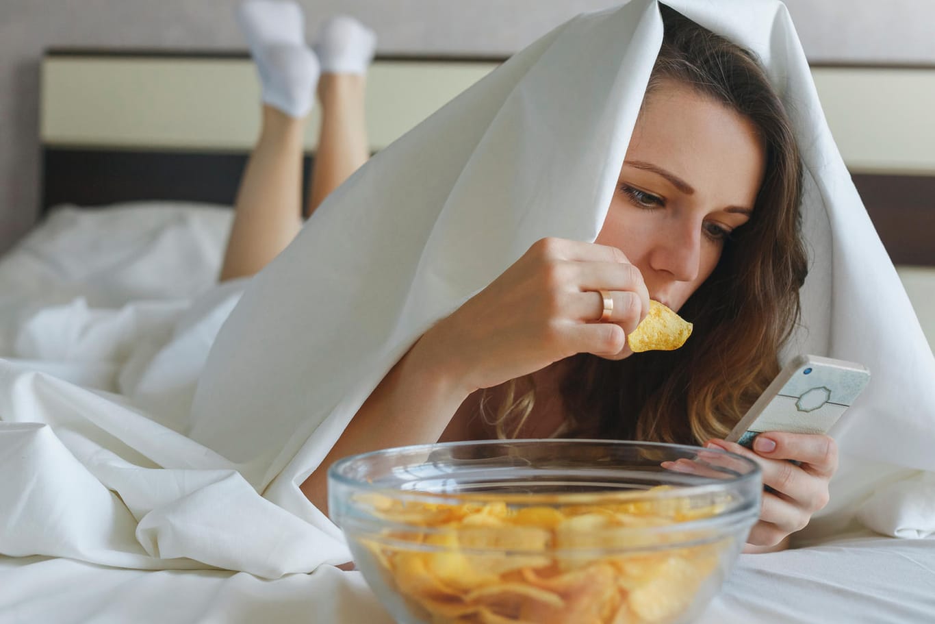 Eine junge Frau isst Chips im Bett und schaut dabei auf ihr Handy. Falsche Ernährungsgewohnheiten können dazu führen, dass man schlechter schläft.