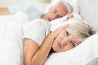 Frau hält ihre Ohren zu, weil der Mann schnarcht. Neben einem Schnarcher zu liegen, kann die Nachtruhe empfindlich stören.