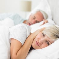 Frau hält ihre Ohren zu, weil der Mann schnarcht. Neben einem Schnarcher zu liegen, kann die Nachtruhe empfindlich stören.
