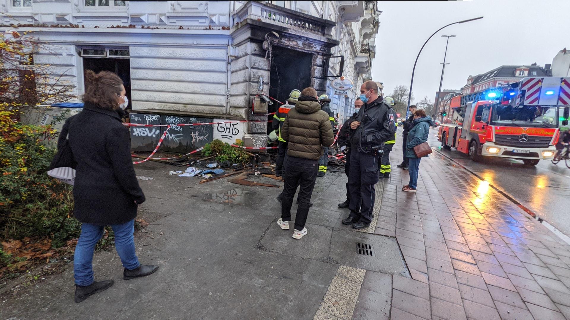 Einsatzkräfte der Feuerwehr stehen vor dem Eingang der Kneipe. Das Feuer soll von brennendem Unrat vor dem Haus übergesprungen sein. Einsatzkräfte der Feuerwehr stehen vor dem Eingang der Kneipe. Das Feuer soll von brennendem Unrat vor dem Haus übergesprungen sein.