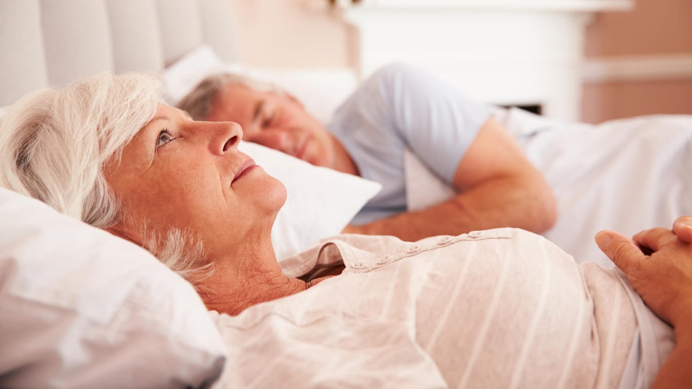 Ältere Dame liegt wach neben ihren Partner im Bett: Im Alter nehmen die Tiefschlafphasen immer mehr ab und der Schlaf wird leichter.