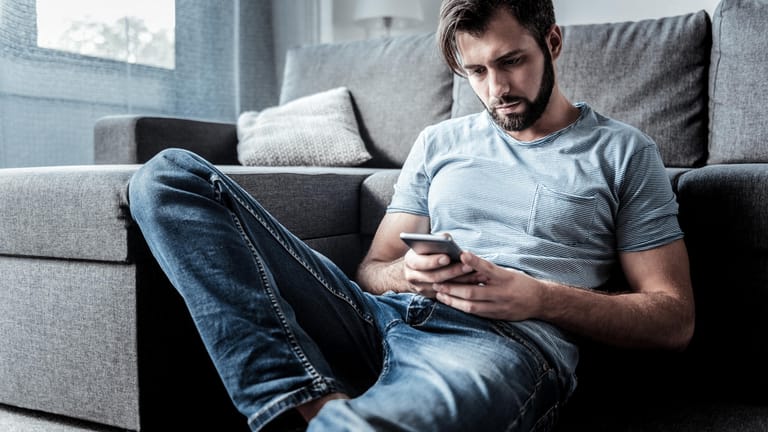 Ein Mann sitzt auf dem Boden an ein Sofa gelehnt und schaut auf sein Smartphone.