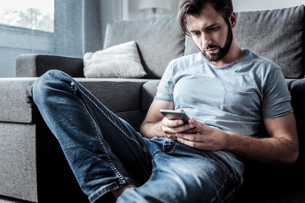 Ein Mann sitzt auf dem Boden an ein Sofa gelehnt und schaut auf sein Smartphone.