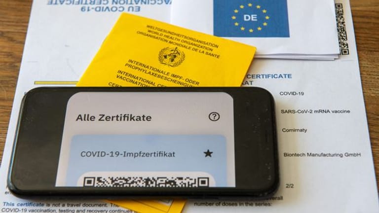 Ein Impfpass und ein Smartphone, auf dem die App CovPass läuft, liegen auf einem Impfzertifikat, das von einer Apotheke ausgestellt wurde.
