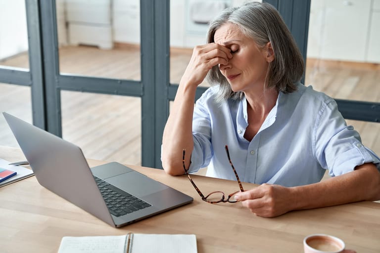 Überarbeitete ältere Frau sitzt am Tisch und fasst sich ins Gesicht: Stress ist die häufigste Ursache für Schlafstörungen. Das ständige Nachdenken über private oder berufliche Probleme hält viele vom Ein- oder Durchschlafen ab.
