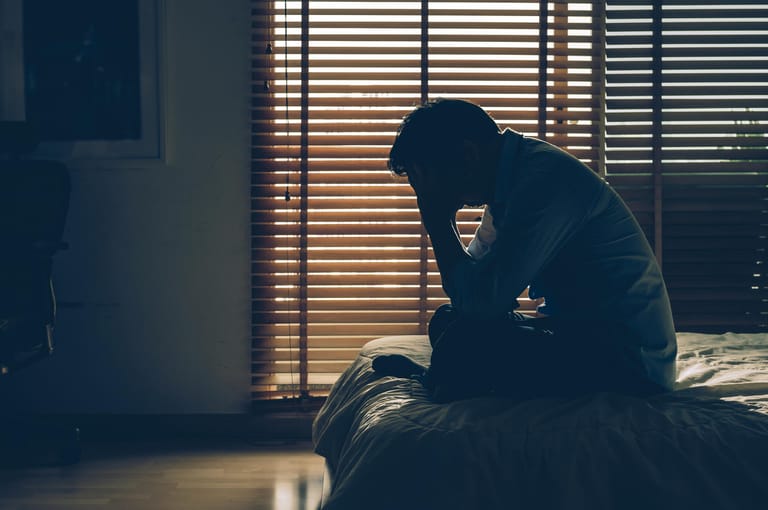 Mann sitzt auf Bett im abgedunkelten Zimmer: Depressionen sind nicht nur tagsüber belastend, sondern stören auch nachts den Schlaf. Vor allem ständige Gedankenkarusselle verhindern ein angenehmes Ein- und Durchschlafen.