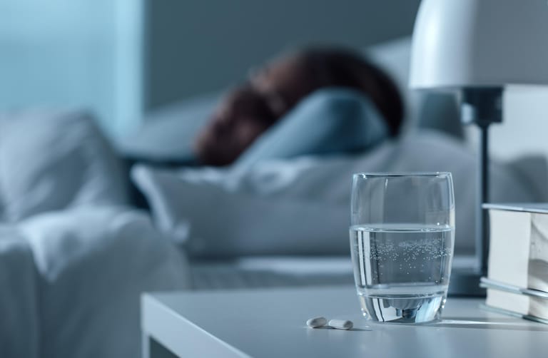 Frau liegt im Bett und auf ihre Nachtisch ein Glas Wasser mit Medikamenten: Medikamente, die die Ausschüttung bestimmter Hormone im Gehirn auslösen, wirken sich häufig negativ auf den Schlaf aus.