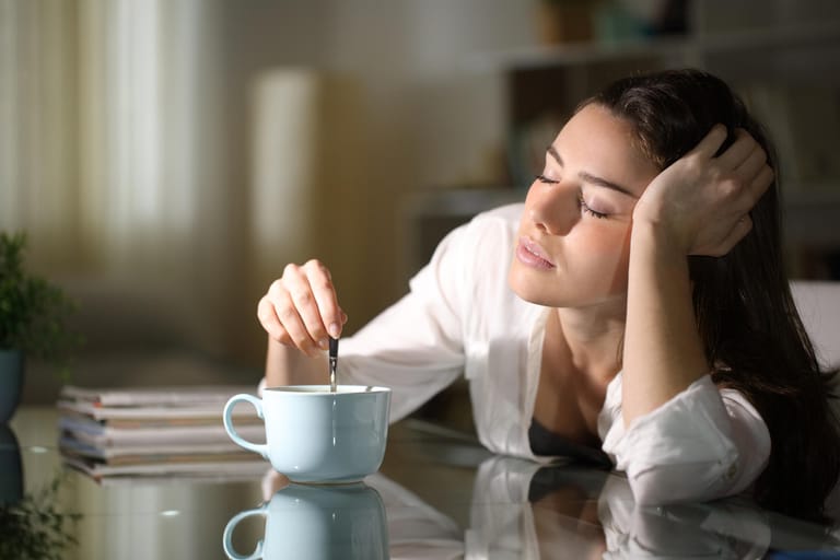 Müde junge Frau trinkt Kaffee: Koffeinhaltige Wachmacher wie Kaffee und Tee, aber auch Alkohol, sollte man nicht vor dem Schlafen gehen trinken. Das Koffein verzögert das Einschlafen und kann die Schlafqualität beinträchtigen. Alkoholkonsum macht zwar müde, doch der Schlaf ist nicht erholsam und wird oft von Wachphasen unterbrochen.