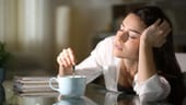 Müde junge Frau trinkt Kaffee: Koffeinhaltige Wachmacher wie Kaffee und Tee, aber auch Alkohol, sollte man nicht vor dem Schlafen gehen trinken. Das Koffein verzögert das Einschlafen und kann die Schlafqualität beinträchtigen. Alkoholkonsum macht zwar müde, doch der Schlaf ist nicht erholsam und wird oft von Wachphasen unterbrochen.