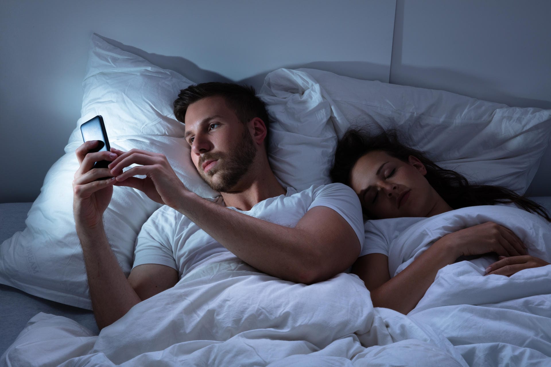 Mann liegt im Bett und benutzt Handy vor dem Schlafen gehen: Kurz vor dem Schlafengehen sollte man nicht mehr h auf das Smartphone oder den Laptop schauen. Denn blauwelliges Licht verhindert die Melatoninausschüttung, so dass man länger wachbleibt und Schwierigkeiten hat einzuschlafen.
