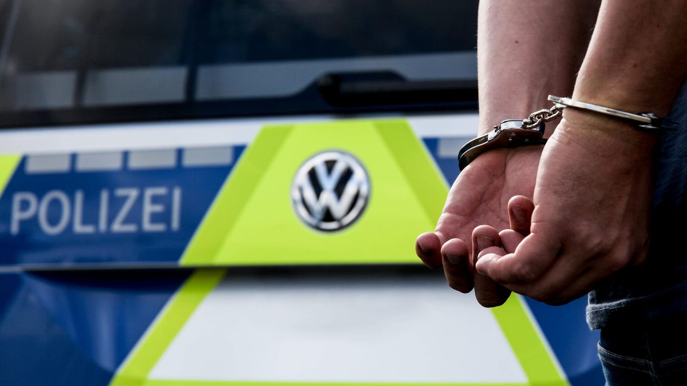 Eine Person steht vor einem Polizeiauto und trägt Handschellen (Symbolbild): Die Tat hat sich bereits im Oktober zugetragen.