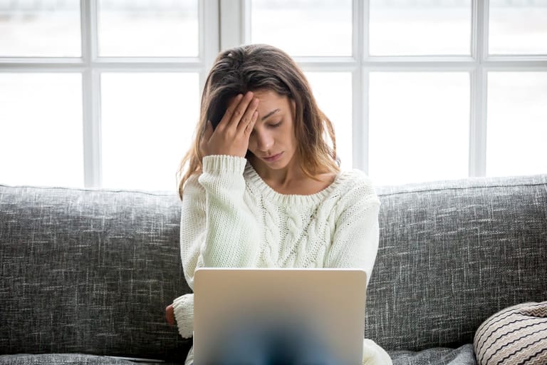 Unter Stress leidende junge Frau sitz mit Laptop auf dem Sofa: Stress: Bei hoher Belastung verbraucht der Körper viel Energie und damit auch Mineralstoffe. Liegt ein Mangel vor, geht er an die körpereignen Reserven. Wenn dabei den Haarwurzeln wichtige Nährstoffe entzogen werden. Kann es zu Haarausfall kommen.