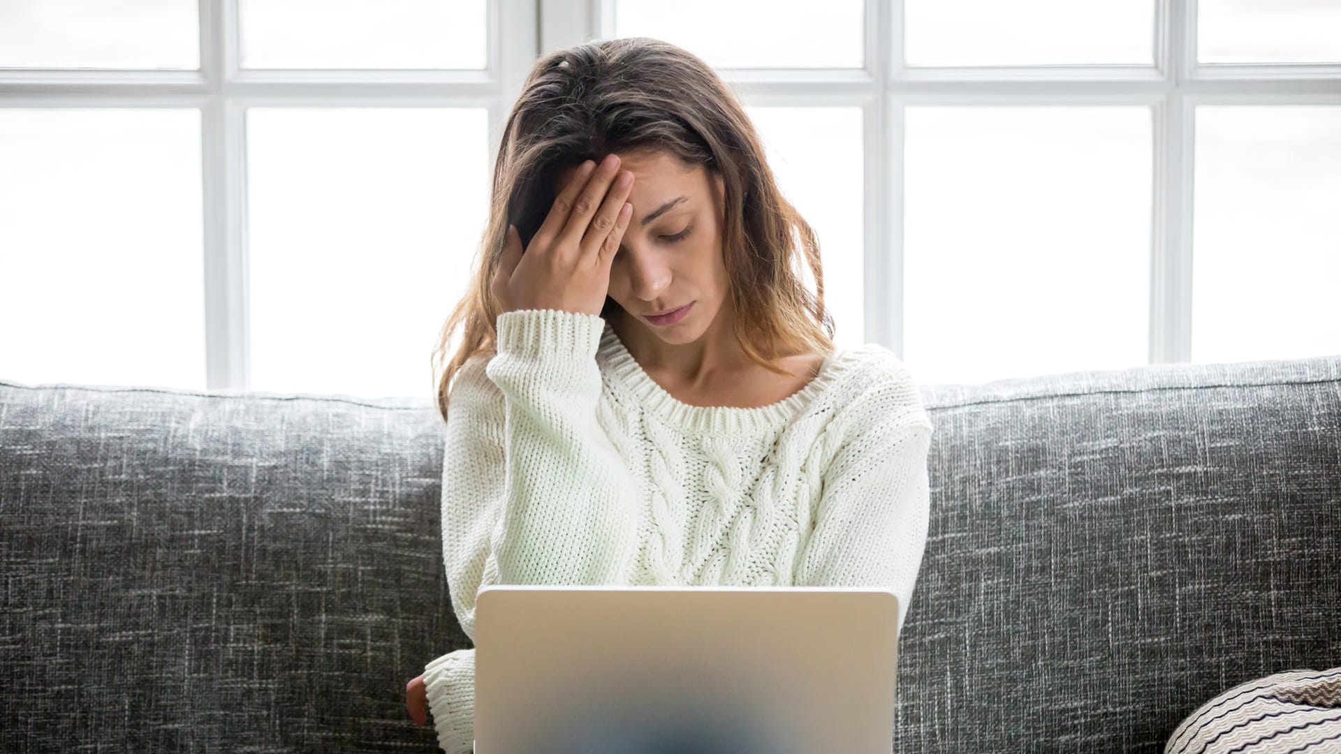 Unter Stress leidende junge Frau sitz mit Laptop auf dem Sofa: Stress: Bei hoher Belastung verbraucht der Körper viel Energie und damit auch Mineralstoffe. Liegt ein Mangel vor, geht er an die körpereignen Reserven. Wenn dabei den Haarwurzeln wichtige Nährstoffe entzogen werden. Kann es zu Haarausfall kommen.