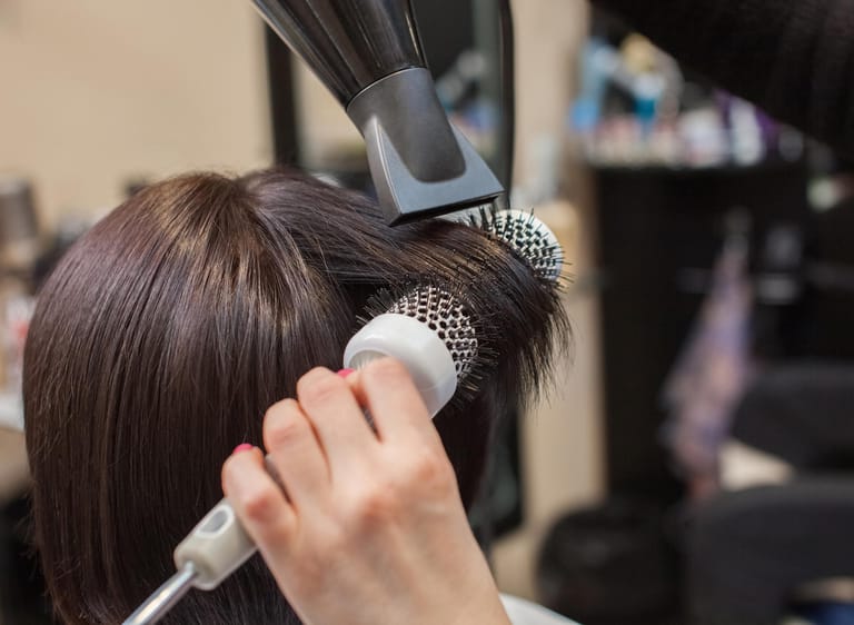 Friseur föhnt Frau die Haare: Zu viel Hitze durch Föhn und Glätteisen, aber auch regelmäßiges Färben, schaden dem Haar. Wer seinen Haaren das zumutet, riskiert dass sie schneller austrocknen und abbrechen.