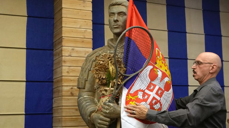 Ein Mann drapiert die serbische Flagge um die Statue des Tennisspielers Djokovic vor der Pressekonferenz.