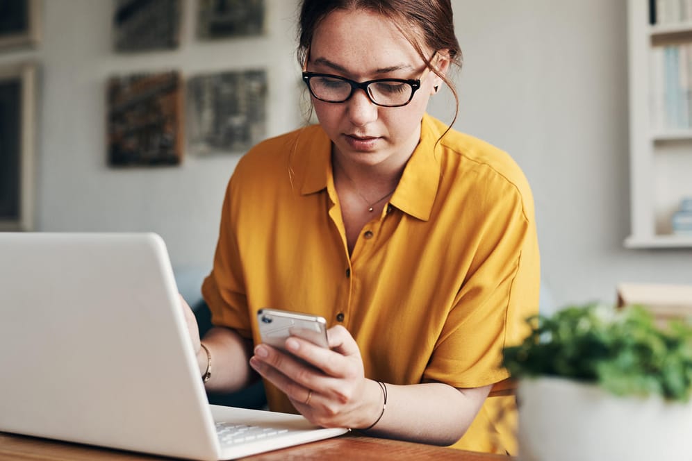 Frau schaut während der Arbeit am Laptop auf ihr Handy: Der Blick auf das Handy gehört zu den häufigsten Ablenkungsfaktoren. Beim Bewältigen wichtiger Aufgaben kann es helfen, das Handy vorübergehend stumm zu stellen.