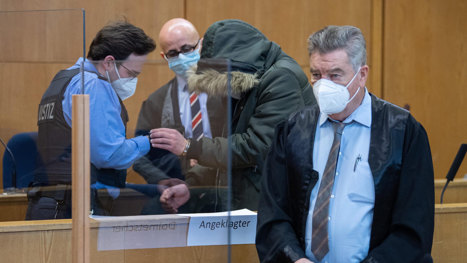 Der Angeklagte zwischen seinen Verteidigern Oussama Al-Agi (2.v.l.) und Ulrich Endres (r).: Ein Justizbeamter nimmt ihm die Handschellen ab.