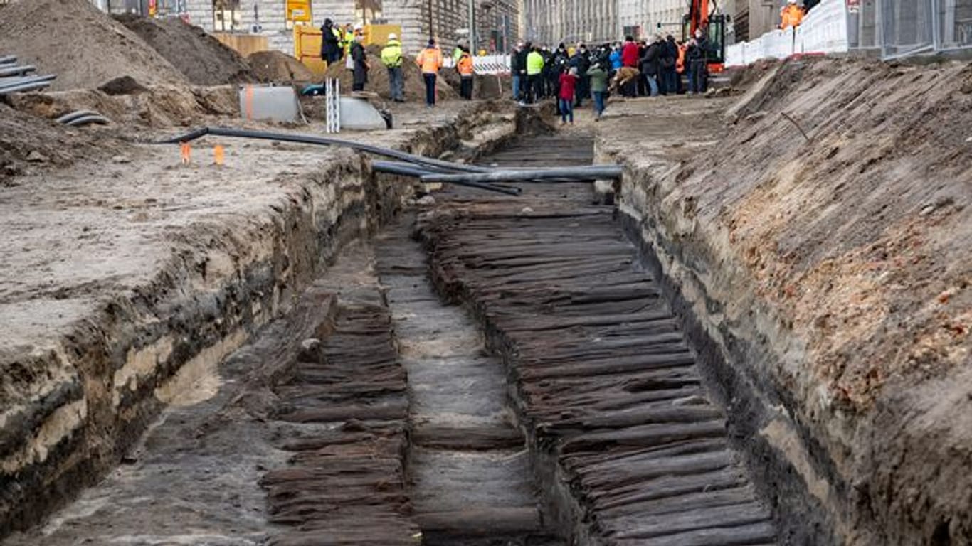 Bei einem Pressetermin stehen zahlreiche Menschen an dem ausgegrabenen mittelalterlichen Bohlendamm, der bei Bauarbeiten unweit des Roten Rathauses entdeckt wurde: Die Straße ist Hunderte Jahre alt.