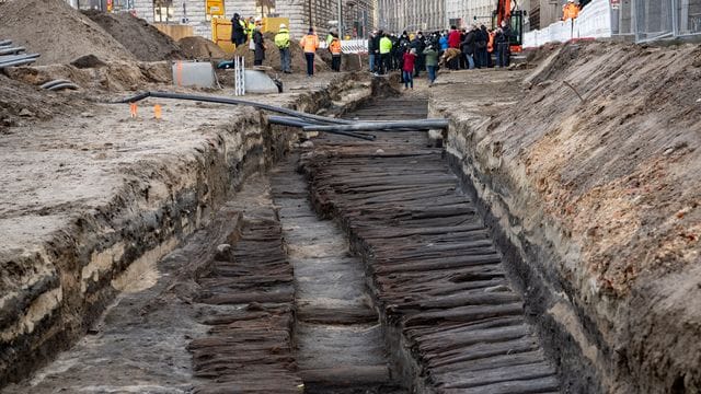 Bei einem Pressetermin stehen zahlreiche Menschen an dem ausgegrabenen mittelalterlichen Bohlendamm, der bei Bauarbeiten unweit des Roten Rathauses entdeckt wurde: Die Straße ist Hunderte Jahre alt.