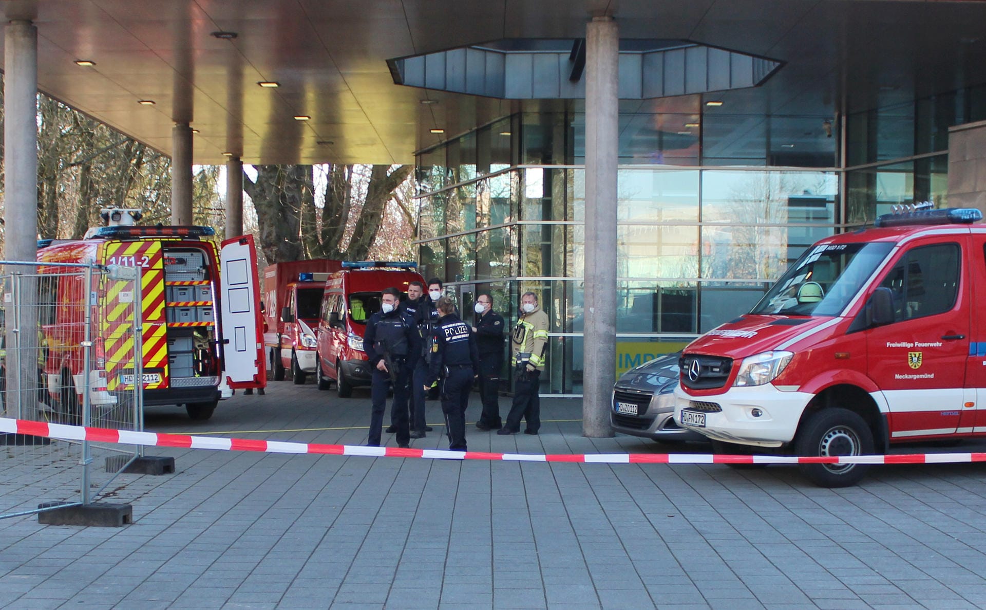 Polizisten stehen neben Krankenwagen und Fahrzeugen der Feuerwehr auf der Gelände der Heidelberger Universität an einem Eingang zu einer Klinik, die sich auf dem Campus befindet. Einer der Polizisten hält dabei eine Maschinenpistole in der Hand.