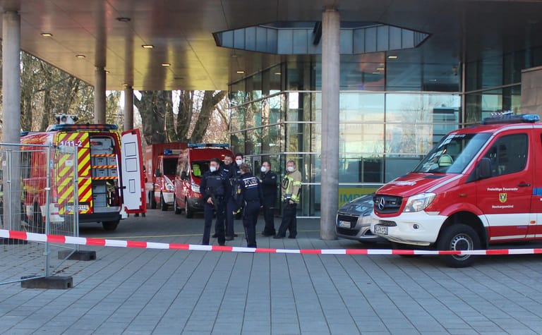 Polizisten stehen neben Krankenwagen und Fahrzeugen der Feuerwehr auf der Gelände der Heidelberger Universität an einem Eingang zu einer Klinik, die sich auf dem Campus befindet. Einer der Polizisten hält dabei eine Maschinenpistole in der Hand.