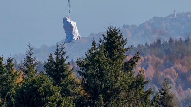 Ein Hubschrauber der italienischen Feuerwehr fliegt die in weiße Planen gehüllte Kabine der abgestürzten Mottarone-Seilbahngondel vom Lago Maggiore ins Tal. Ein Hubschrauber der italienischen Feuerwehr fliegt die in weiße Planen gehüllte Kabine der abgestürzten Mottarone-Seilbahngondel vom Lago Maggiore ins Tal.