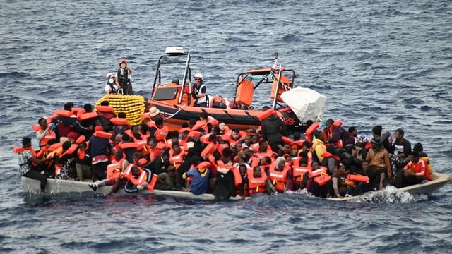 Ein Rettungsboot schwimmt hinter einem völlig überfüllten Flüchtlingsboot.