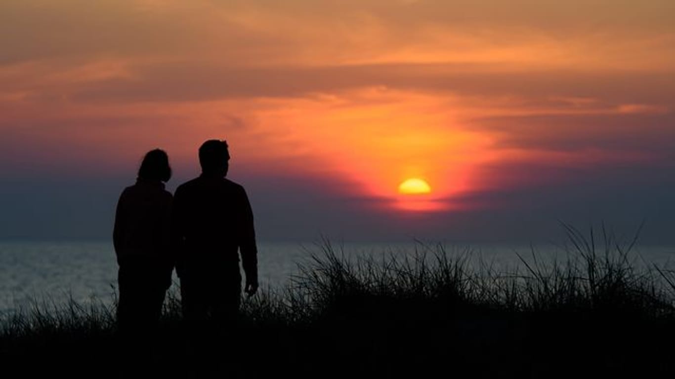 Ein Paar steht während des rötlich-orangenen Sonnenunterganges über der Ostsee an einer Düne am Strand.