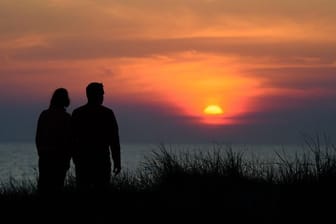Ein Paar steht während des rötlich-orangenen Sonnenunterganges über der Ostsee an einer Düne am Strand.