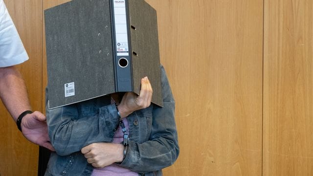 Die Angeklagte verdeckt ihr Gesicht mit einem Aktenordner, während sie in Handschellen in den Schwurgerichtssaal des Landgerichts in Ulm geführt wird.